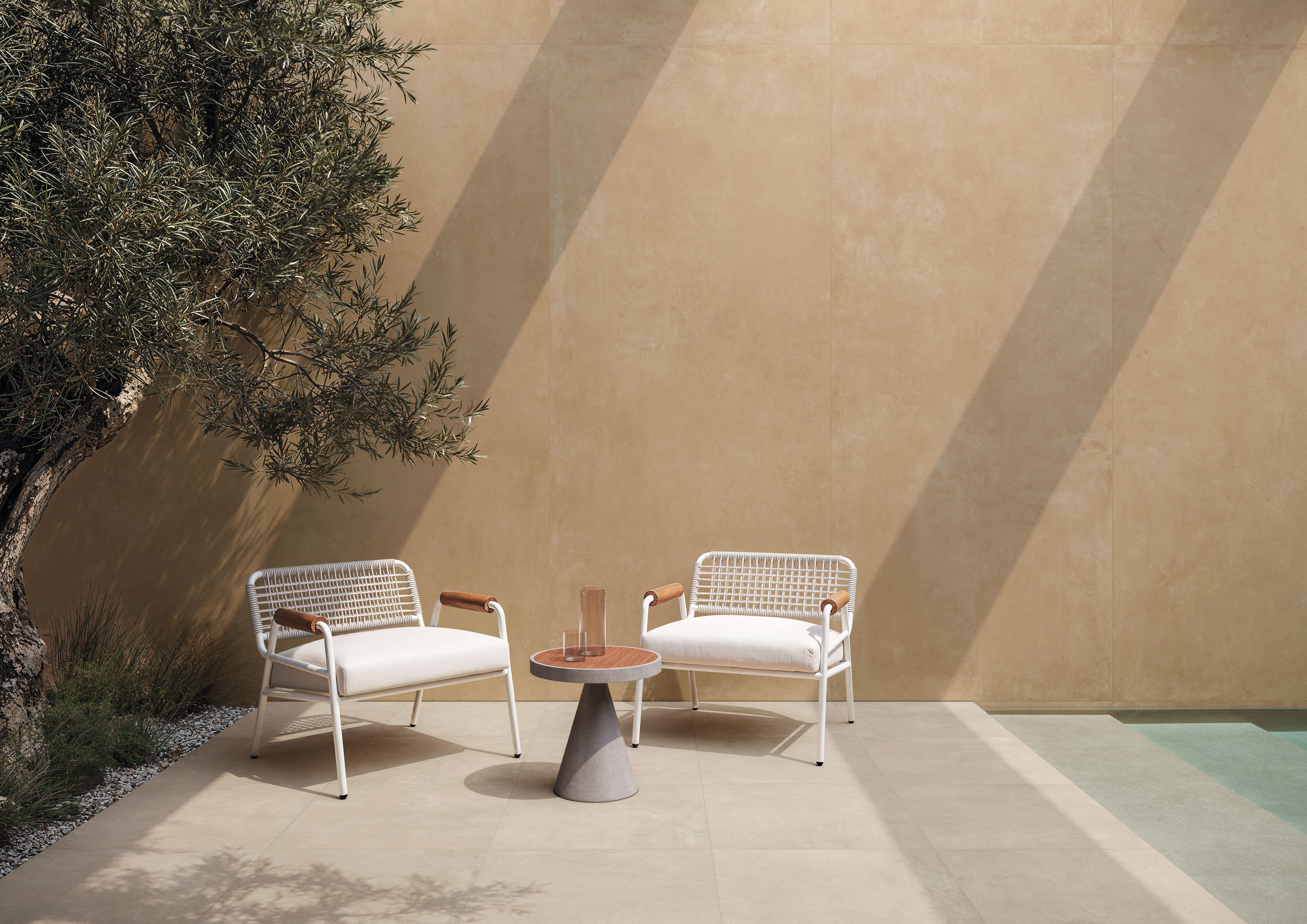 Concrete porcelain tile in Sand on a sunlit courtyard wall, with Beige paving underfoot, an olive tree and a pool edge.