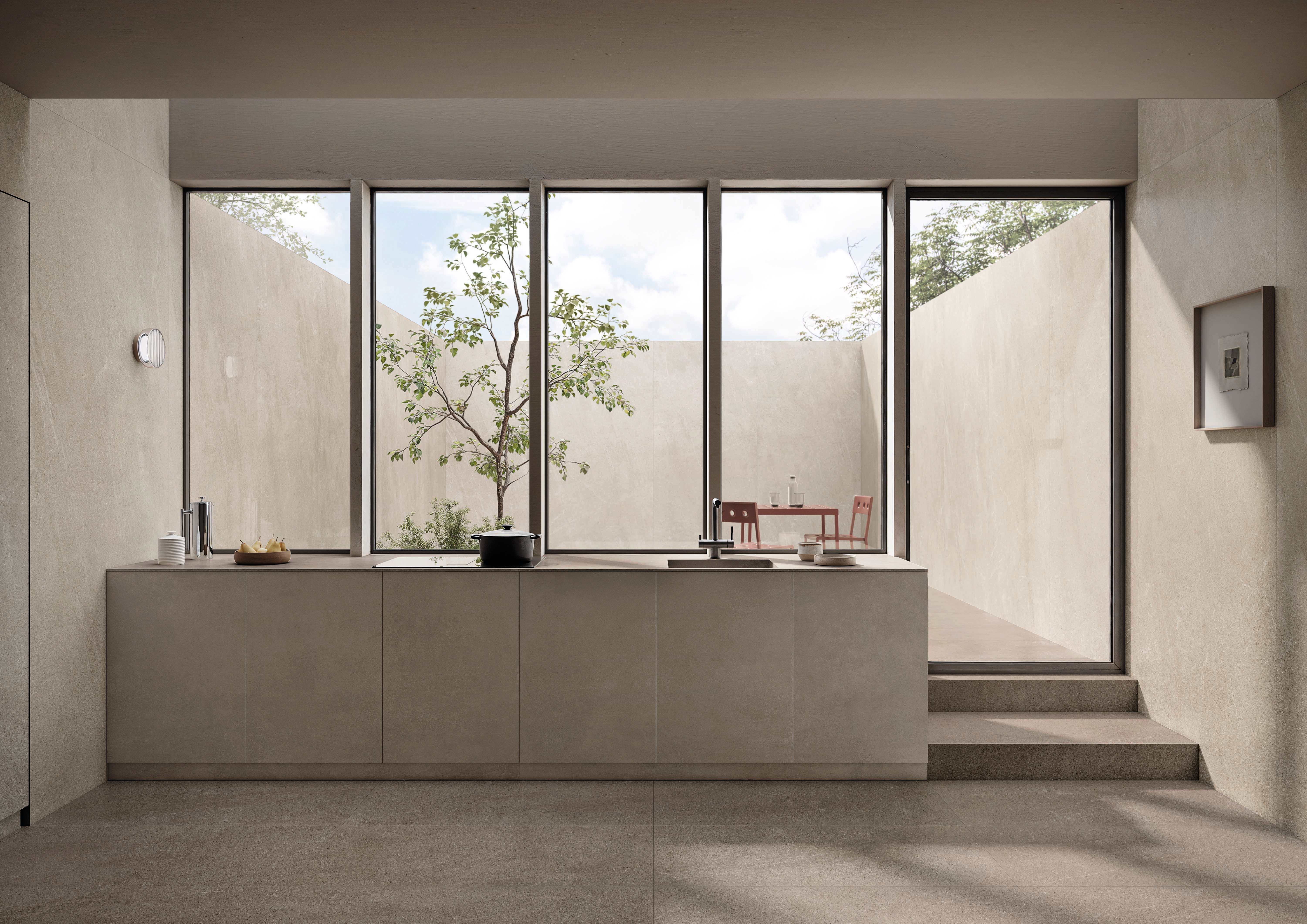 Minimalist open kitchen in pale, warm-toned stone-look Natural porcelain, with a long low island, soft grey floor, full-height courtyard windows and a tree.