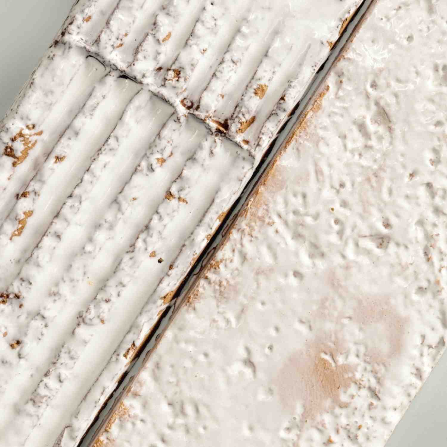 Mutina Chamotte Bianco high-gloss glazed ceramic wall tile extreme close-up showing white cream surface with tactile texture revealing raw clay brown spots and irregular surface relief characteristic of handmade artisan finish
