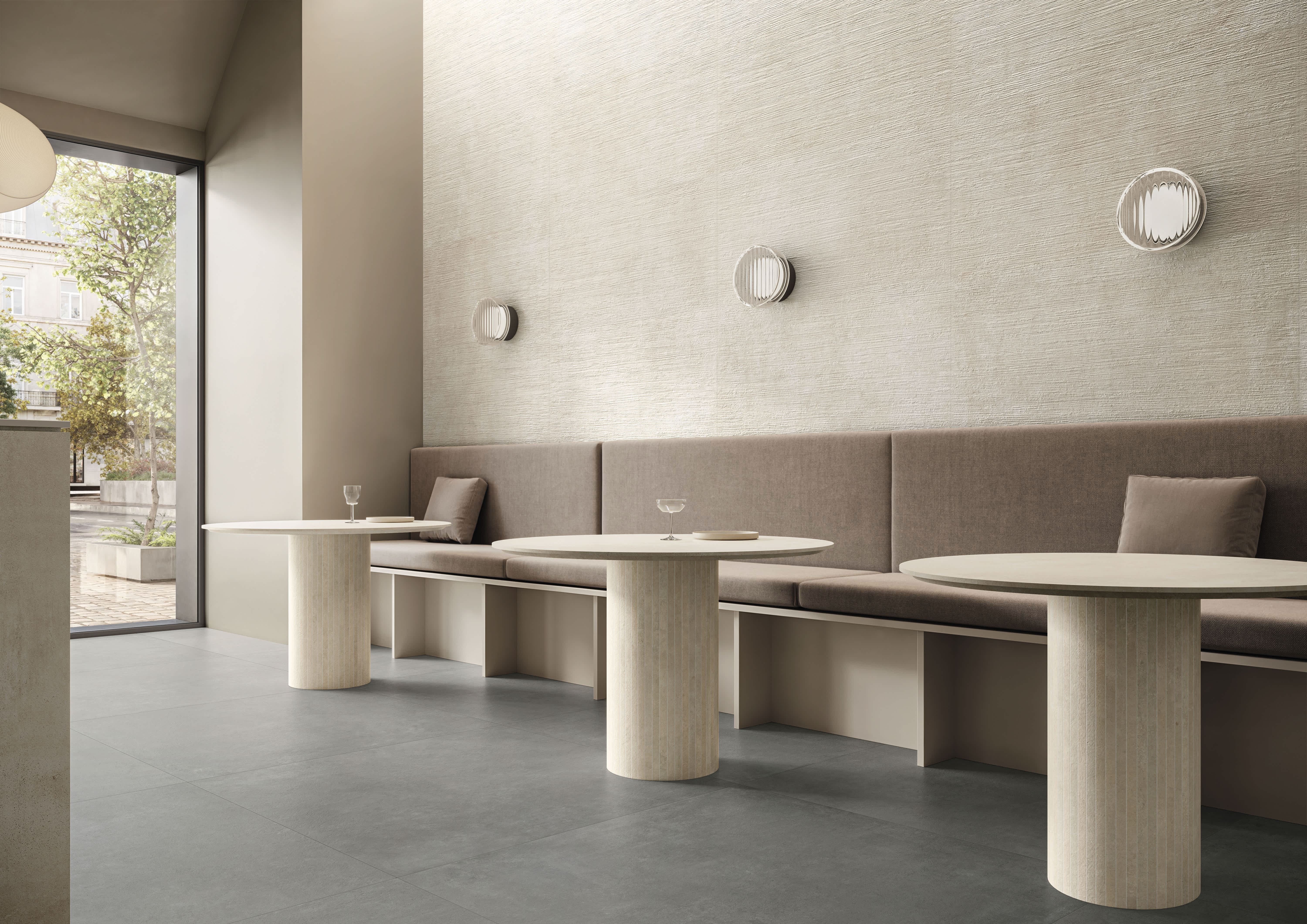 Concrete porcelain tile in a bright café, warm off-white walls above a cool grey floor, with pedestal tables, a long banquette and street-view windows.