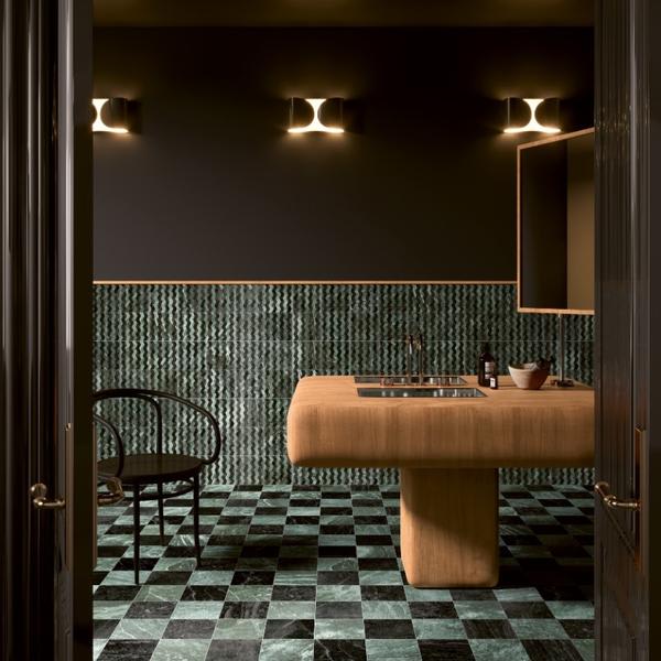 41zero42 Dandy glazed porcelain stoneware dark green chevron wall tiles and green black checkered floor tiles in moody luxury vanity area with brass wall sconces wooden countertop bowl bath accessories and bentwood chair