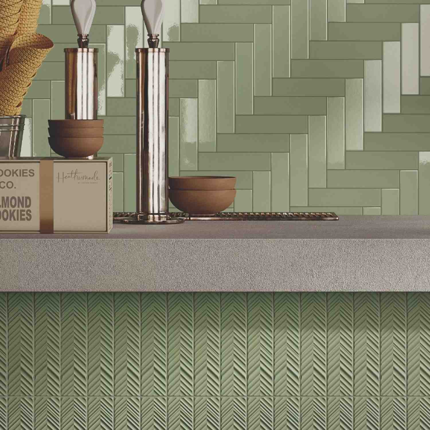 Elios Dust matte chevron ceramic wall tiles in ink sage olive green with mixed chevron pattern layout installed as kitchen backsplash with concrete countertop stacked brown ceramic bowls chrome salt and pepper mills and darker green chevron accent tiles b