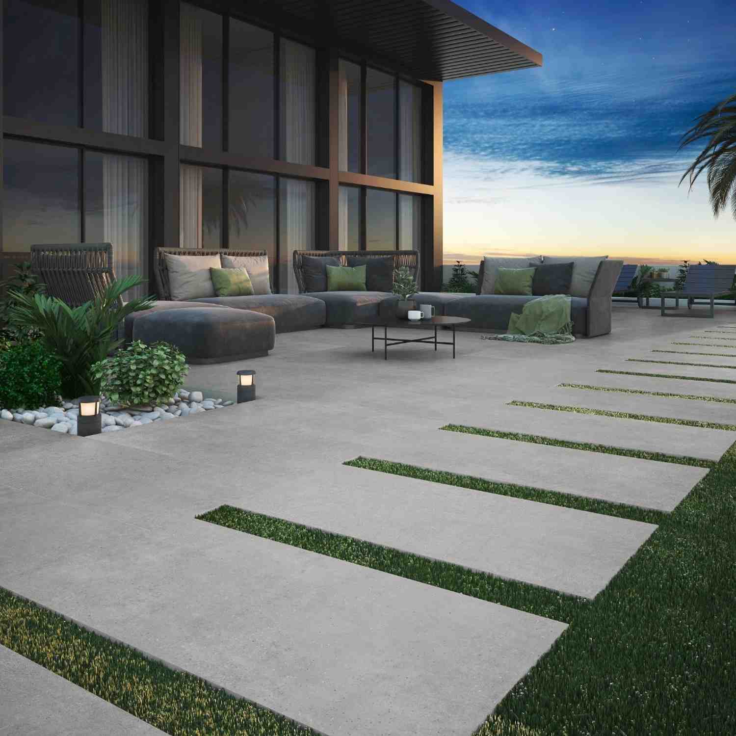 Argenta Gravel porcelain stoneware outdoor patio tiles in grey with stone-cement matte finish on modern terrace at sunset featuring sectional lounge seating with green cushions glass coffee table garden pathway lanterns white pebble accents and floor-to-c