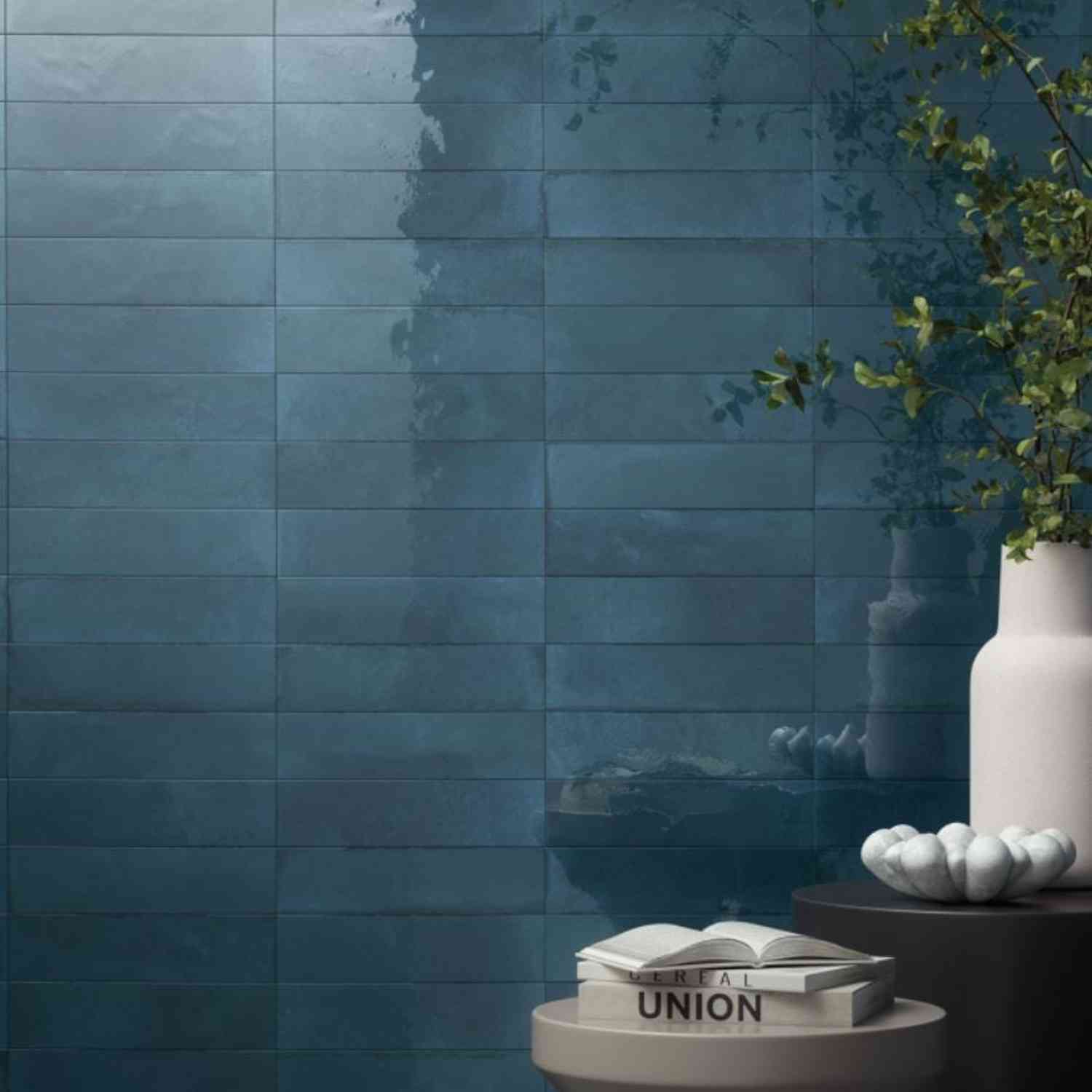 Refin Halo porcelain wall tiles in ink gloss lustre finish with deep blue tone featuring 6x24 format brick-lay pattern with white ceramic vase trailing greenery and subtle tonal variations across the glossy surface