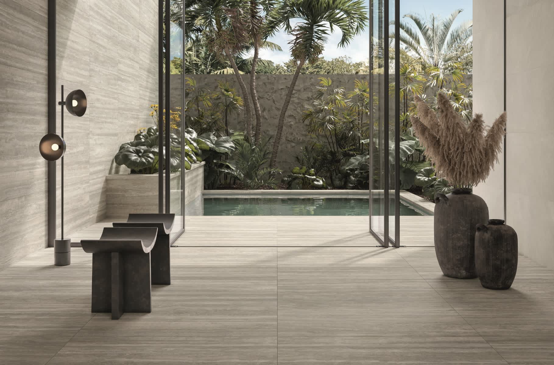 Caesar Iconica mosaic porcelain tile in warm beige tone as flooring in tropical outdoor living area with glass walls overlooking swimming pool and lush garden dark sculptural stools pampas grass vase and globe wall sconces