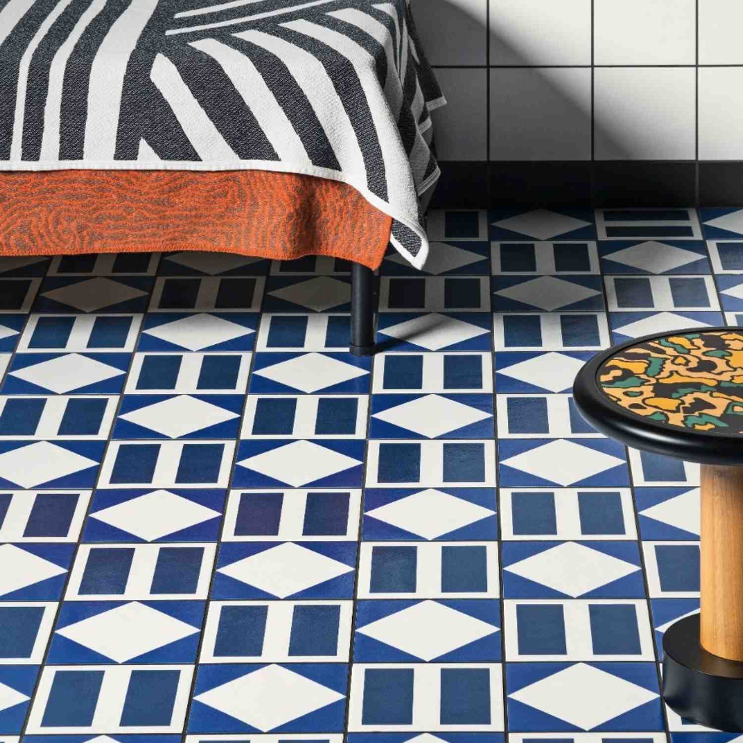 Mutina Margherita blue and white Rhombus diamond pattern porcelain floor tiles in bedroom setting with black and white striped bedding, orange throw, and decorative side table