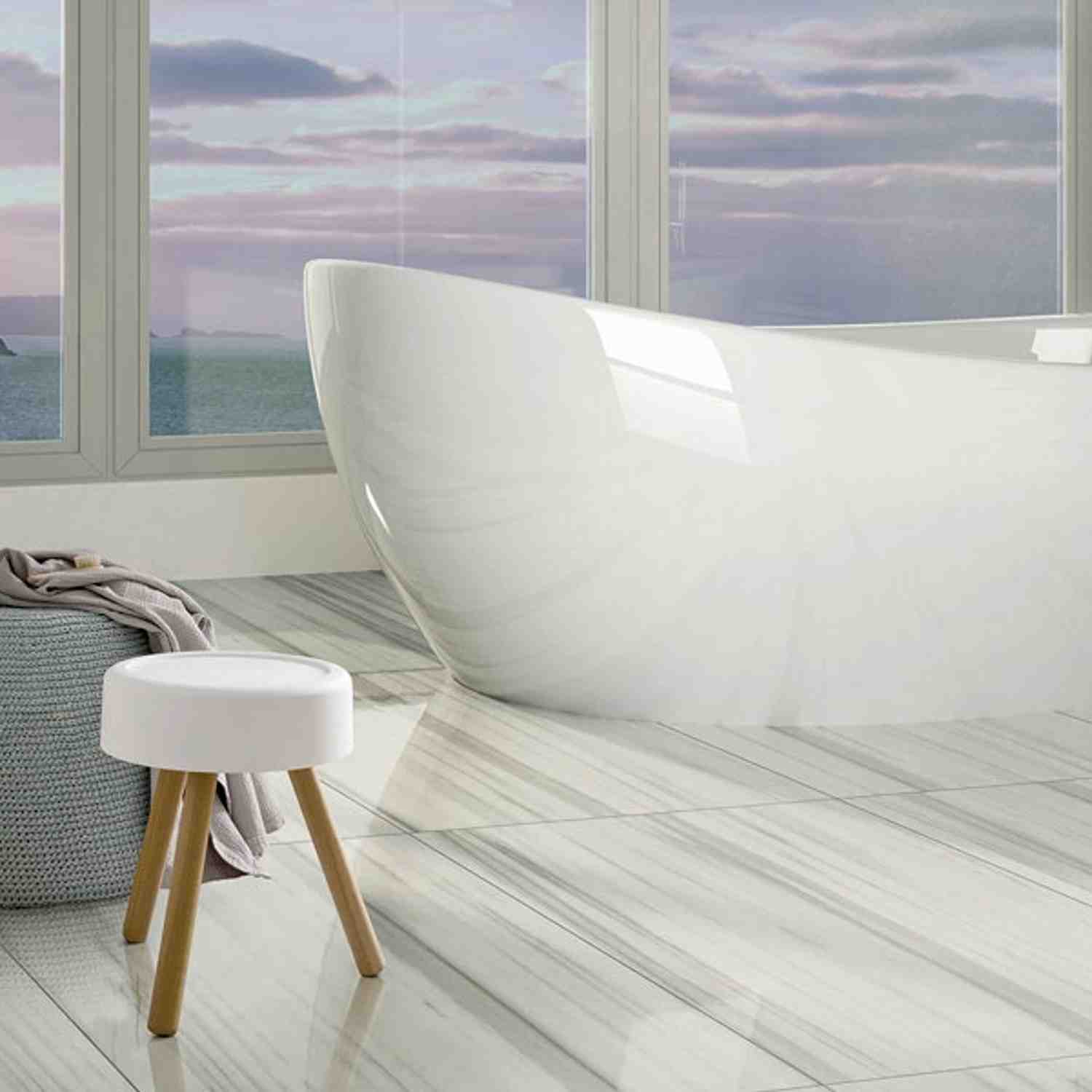 Casalgrande Marmoker Calacatta light marble-look porcelain floor tiles in bright coastal bathroom with freestanding white bathtub, ocean view windows, and minimalist white stool