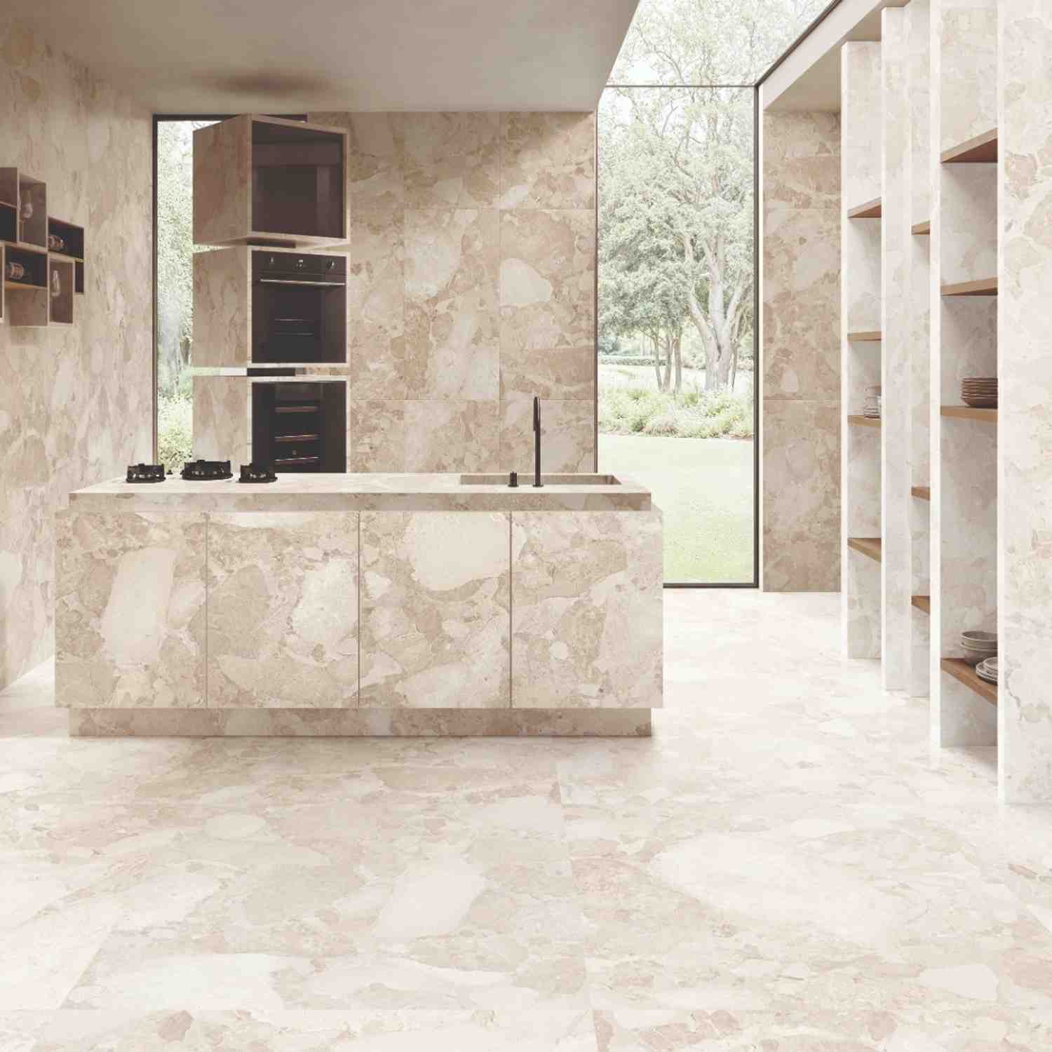 Ergon Materastone Sassi Beige porcelain stoneware wall and floor tiles in a modern kitchen with built-in black ovens, stone island with dark fixtures, open wood shelving, and floor-to-ceiling windows overlooking a garden