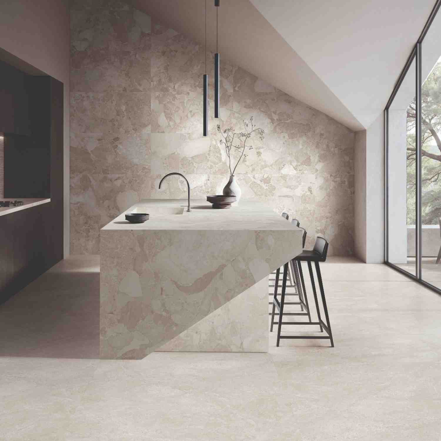 Ergon Materastone Sassi Beige porcelain stoneware walls and floor in a minimalist kitchen with monolithic stone island, black faucet, cylindrical pendant lights, ceramic vase with branches, and angled floor-to-ceiling windows