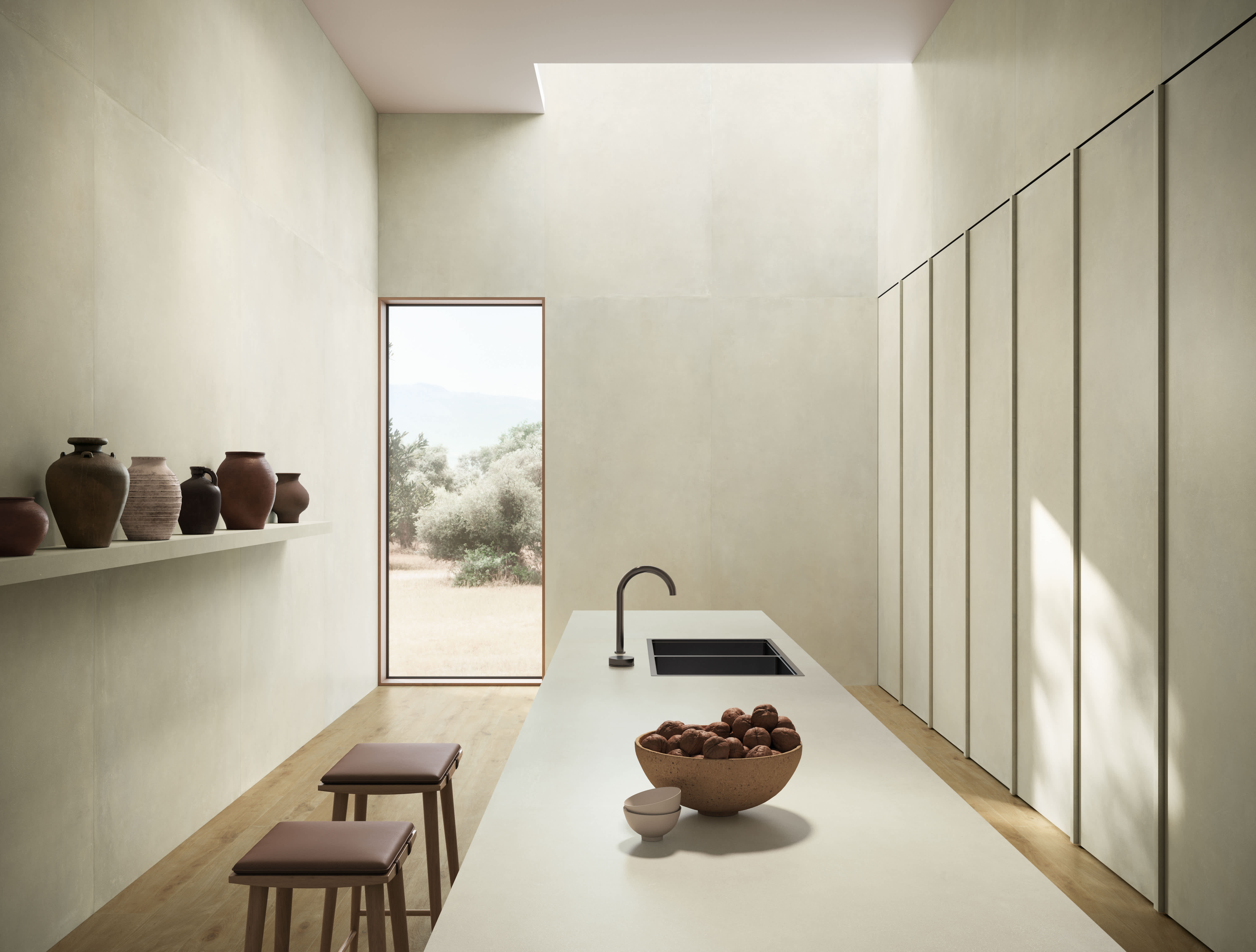 Long galley kitchen in warm off-white I Cementi porcelain slab, with a pale island, terracotta vases and a tall window onto an olive grove.