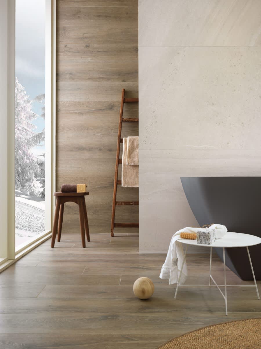 Impronta Nordic Stone Islanda mosaic tiles on bathroom wall with wooden ladder towel rack, stool, dark freestanding bathtub, and snowy mountain view through window