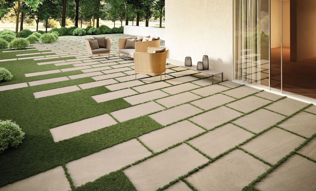Impronta Nordic Stone mosaic porcelain tiles used as outdoor garden pavers with grass joints, styled with wicker lounge furniture, decorative lanterns, and lush green landscaping