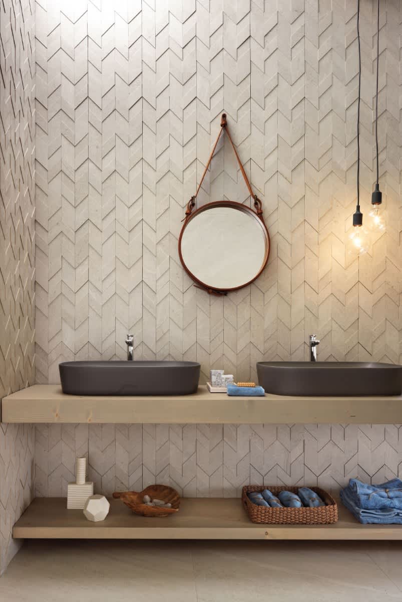 Impronta Nordic Stone mosaic tiles in chevron herringbone wall pattern in a modern bathroom with round leather-strap mirror, Edison bulb pendants, and dark vessel sinks