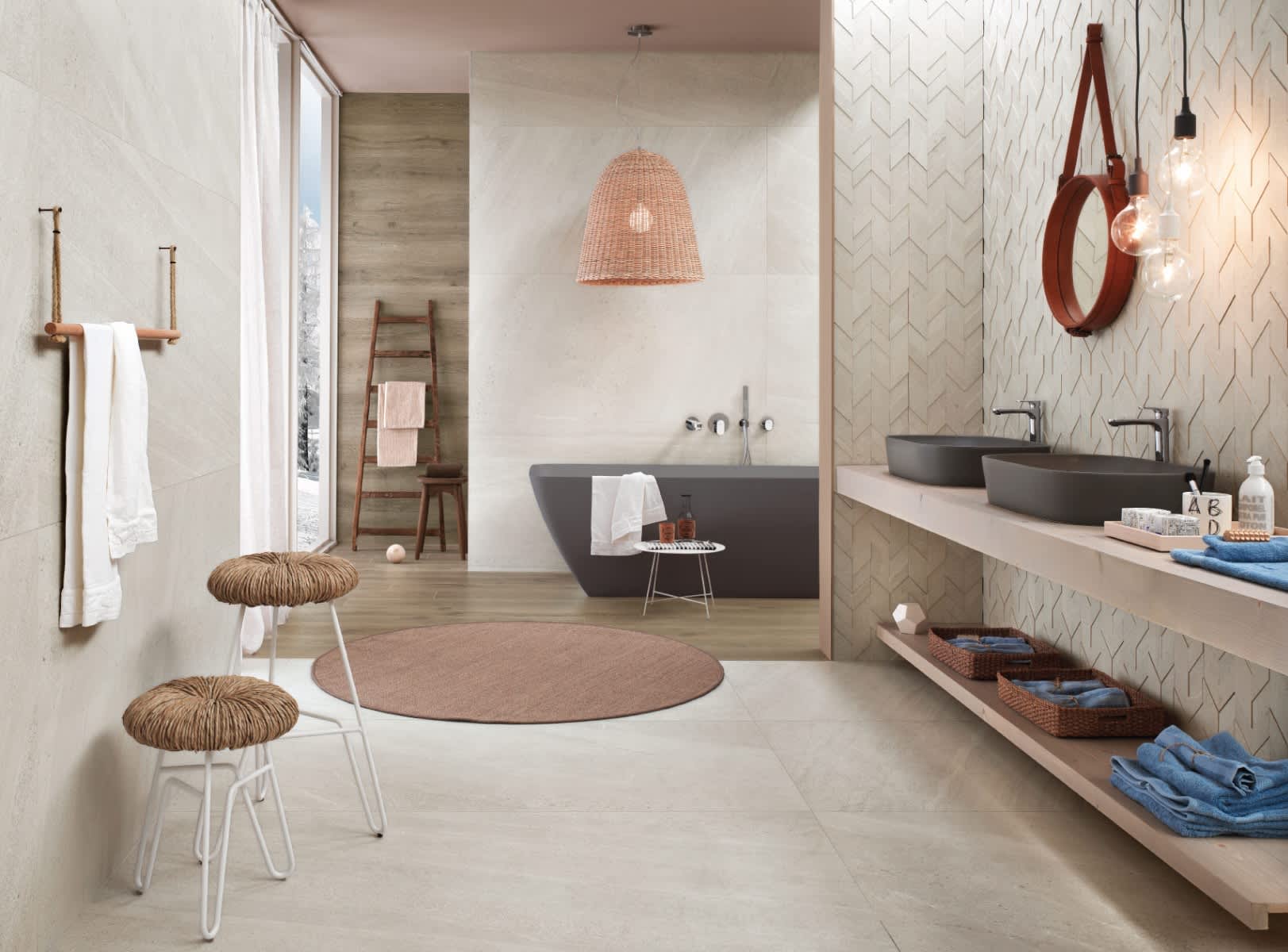 Impronta Nordic Stone Islanda mosaic tiles in chevron pattern on shower wall in a luxury bathroom with dark freestanding bathtub, woven rattan pendant, wooden ladder, round mirror, and floating vanity with vessel sinks