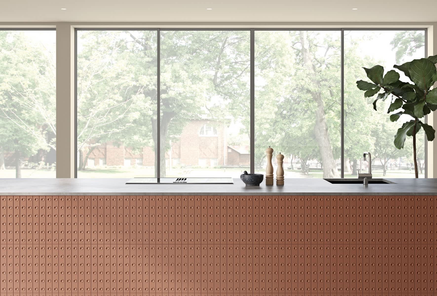 Mutina Punto glazed ceramic tiles in Brun terracotta with raised dot texture on kitchen island front panel, modern kitchen with large windows, dark countertop with cooking accessories and fiddle leaf fig plant
