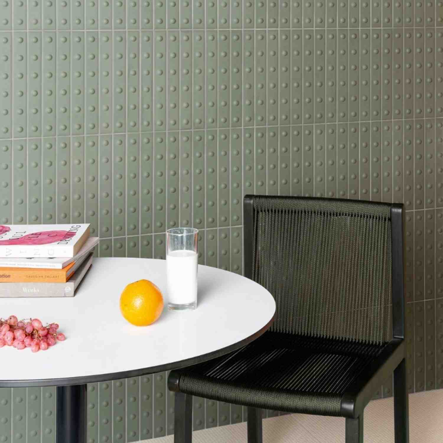 Mutina Punto glazed ceramic wall tiles in Vert green matte with raised dot texture, café-style setting with round white table, books, glass of milk, orange, and dark woven chair