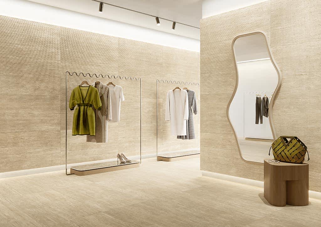 Ceramica Fioranese Senzatempo Trav porcelain stoneware tiles in Beige travertine-look on walls and floor of modern retail boutique with glass clothing racks, wavy contemporary mirror, wooden pedestal with woven basket, and track lighting in commercial int