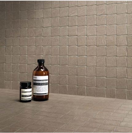 Ergon Stonetalk mosaic porcelain stoneware tiles in taupe matte finish close-up detail showing brick-pattern wall and floor tiles with amber glass bath product bottles as styling accessories
