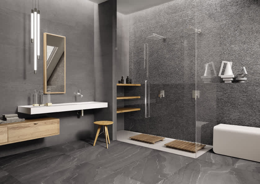 Ergon Stonetalk mosaic porcelain stoneware tiles in Dark Martellata textured finish on bathroom walls and Dark floor with frameless glass walk-in shower, floating white vanity with wooden mirror frame, open wooden shelving, teak bath mat, wooden stool, an