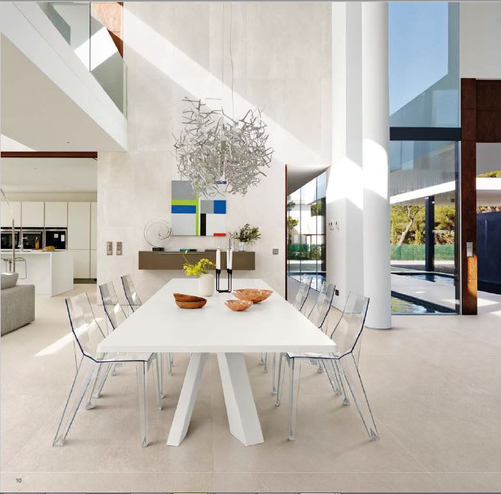 Ergon Stonetalk mosaic porcelain stoneware floor tiles in Sand Minimal light beige finish in contemporary double-height open-plan dining room with white dining table, transparent acrylic chairs, sculptural metal chandelier, wooden bowls, and floor-to-ceil
