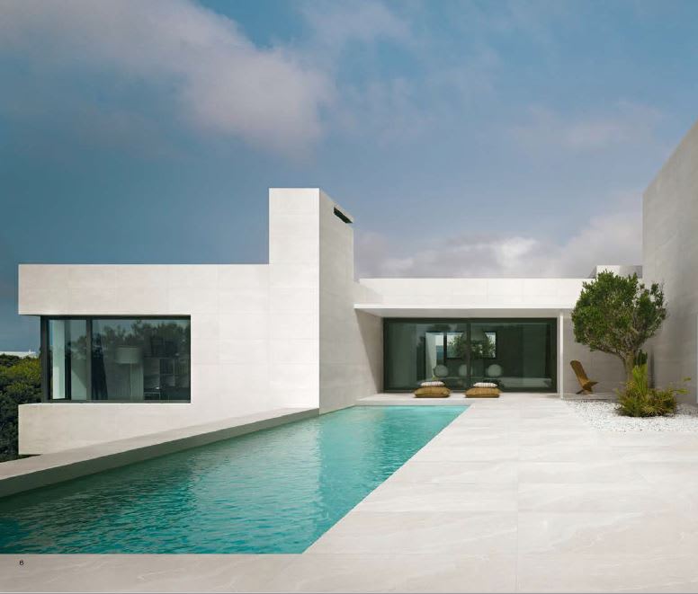 Ergon Stonetalk mosaic porcelain stoneware outdoor paving tiles in White Minimal finish as pool deck surround at contemporary minimalist white villa with turquoise infinity pool, lounge chairs, large glass windows, olive tree, and blue sky