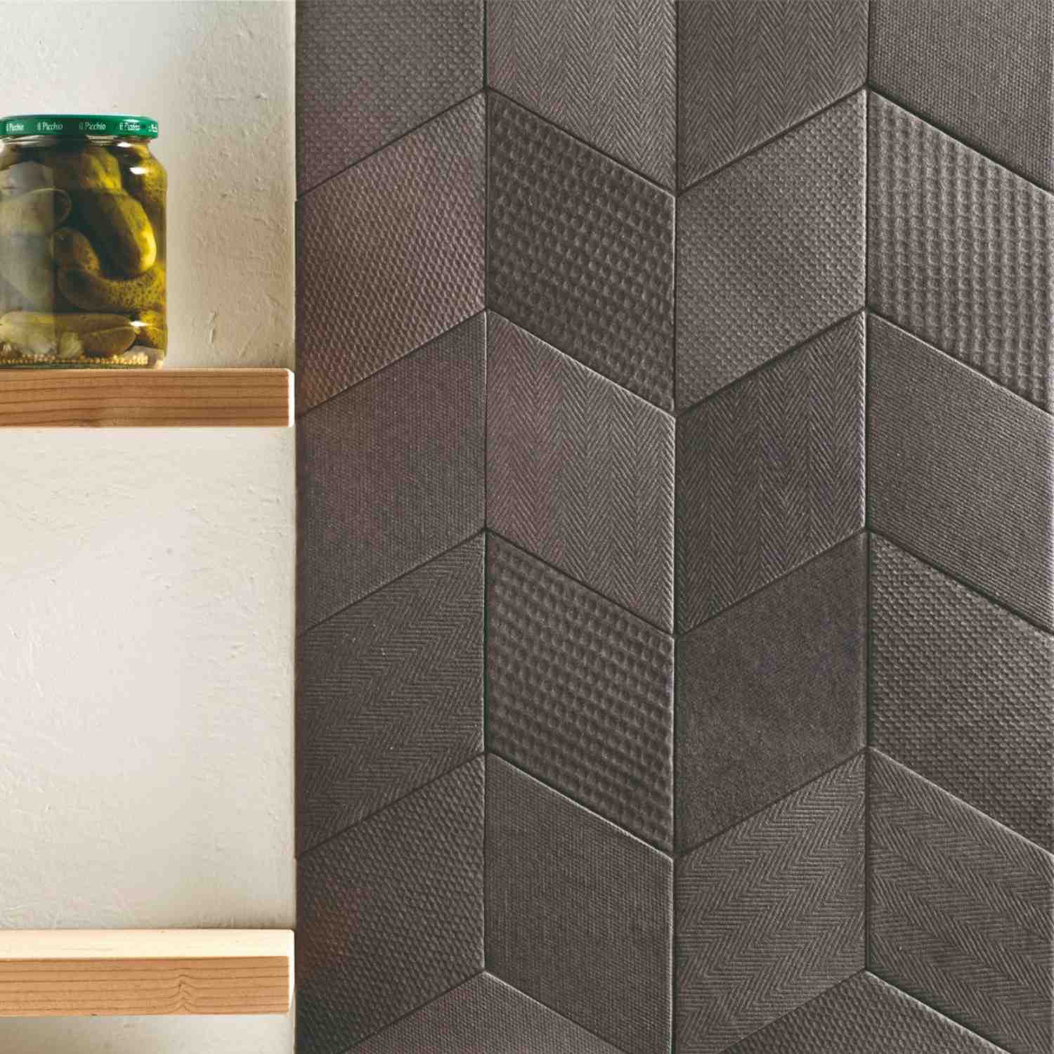 Mutina Tex ceramic wall tile kitchen detail with dark grey three-dimensional textured tiles in herringbone and chevron patterns, wooden shelf with glass jar, and cream wall accent