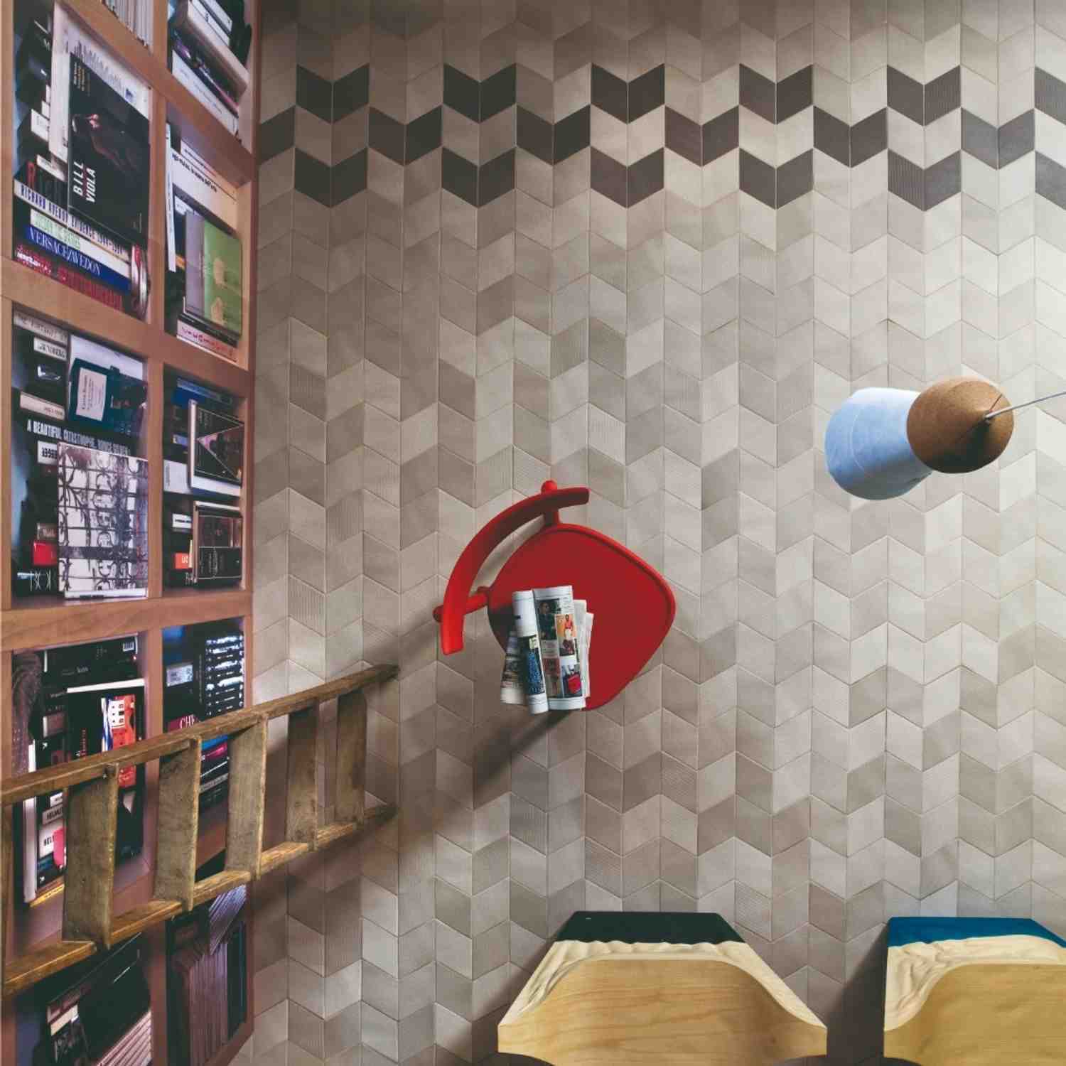 Mutina Tex ceramic wall tile reading nook with beige three-dimensional chevron textured tiles, red accent chair with magazines, wooden bookshelf, and hanging blue and brass pendant light