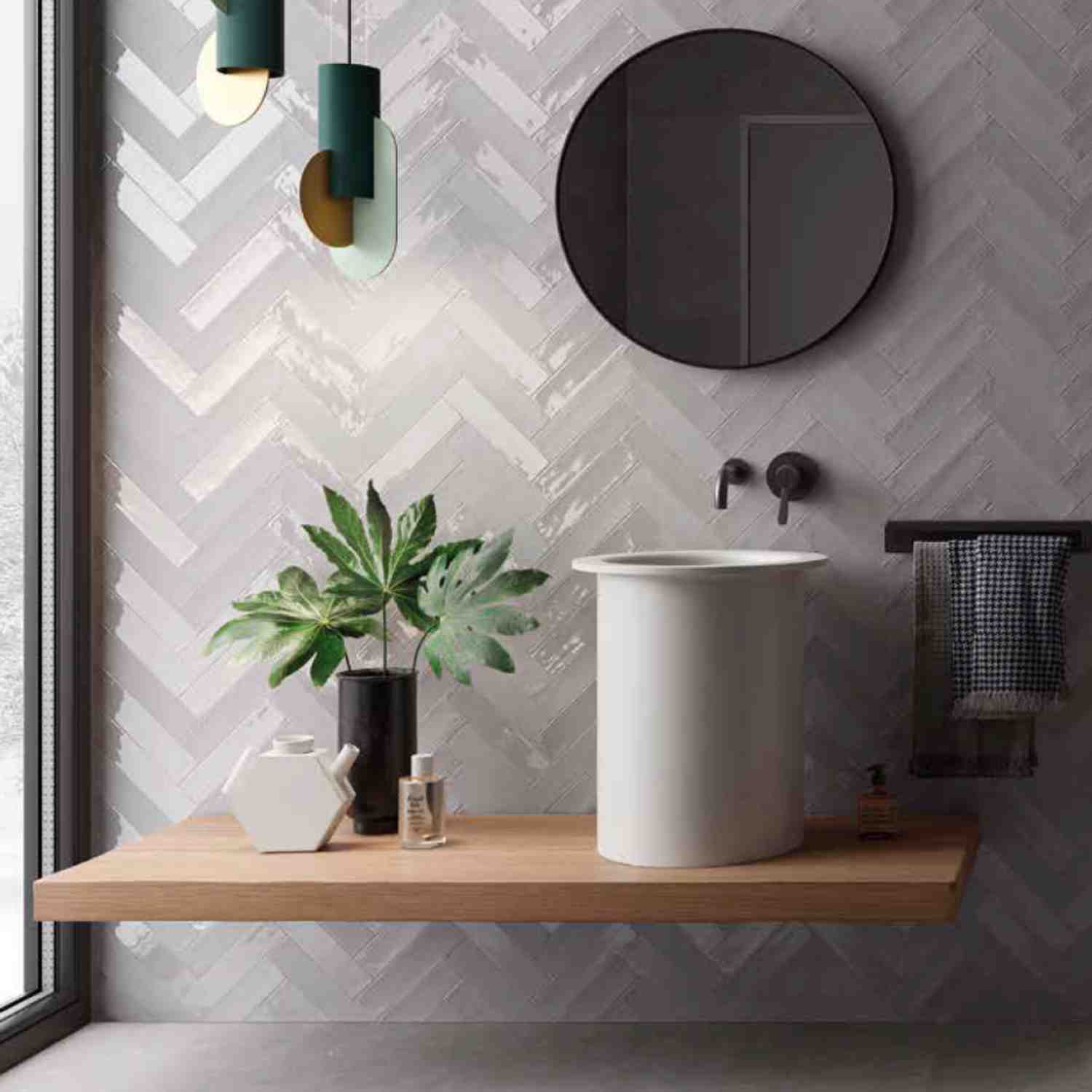 Ergon Total Brick ceramic wall tiles in light grey herringbone pattern in a modern bathroom with round mirror, white pedestal sink, wooden floating shelf, and green plants