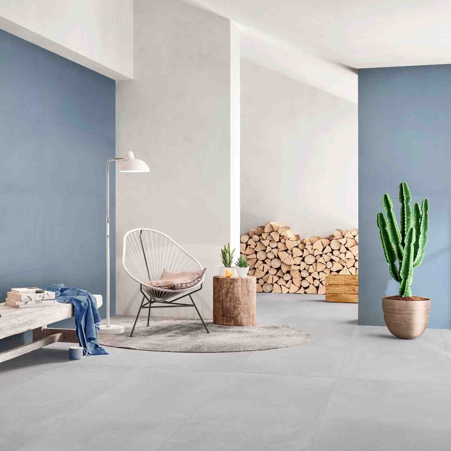 Florina Urban Cement porcelain floor tiles in light grey in a modern living room with blue accent wall, white wire chair, stacked firewood, and cactus plant
