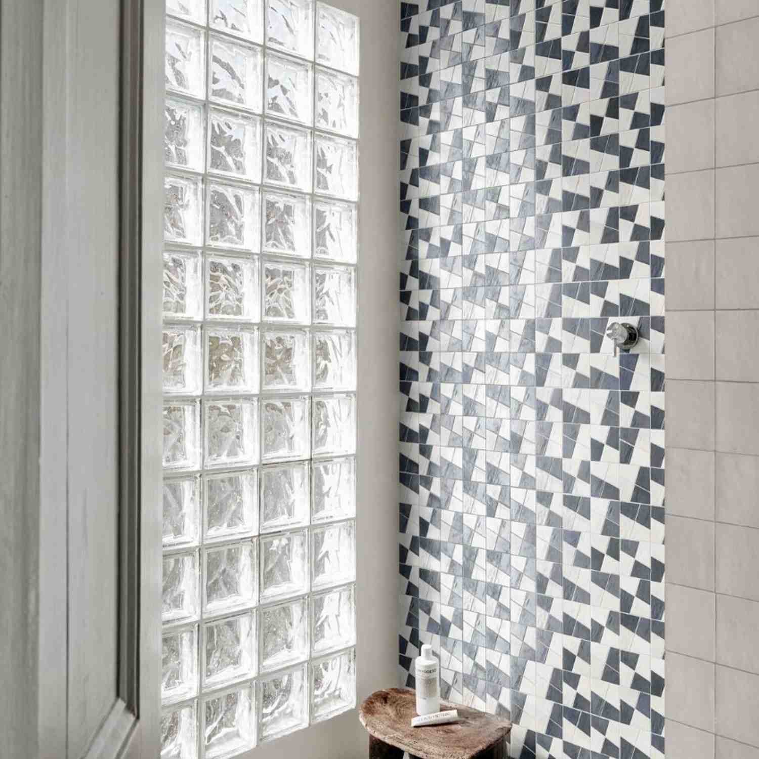 Marazzi Zellige ceramic wall tiles in blue and white geometric chevron pattern in bathroom with glass block window, wooden stool, and white fixtures
