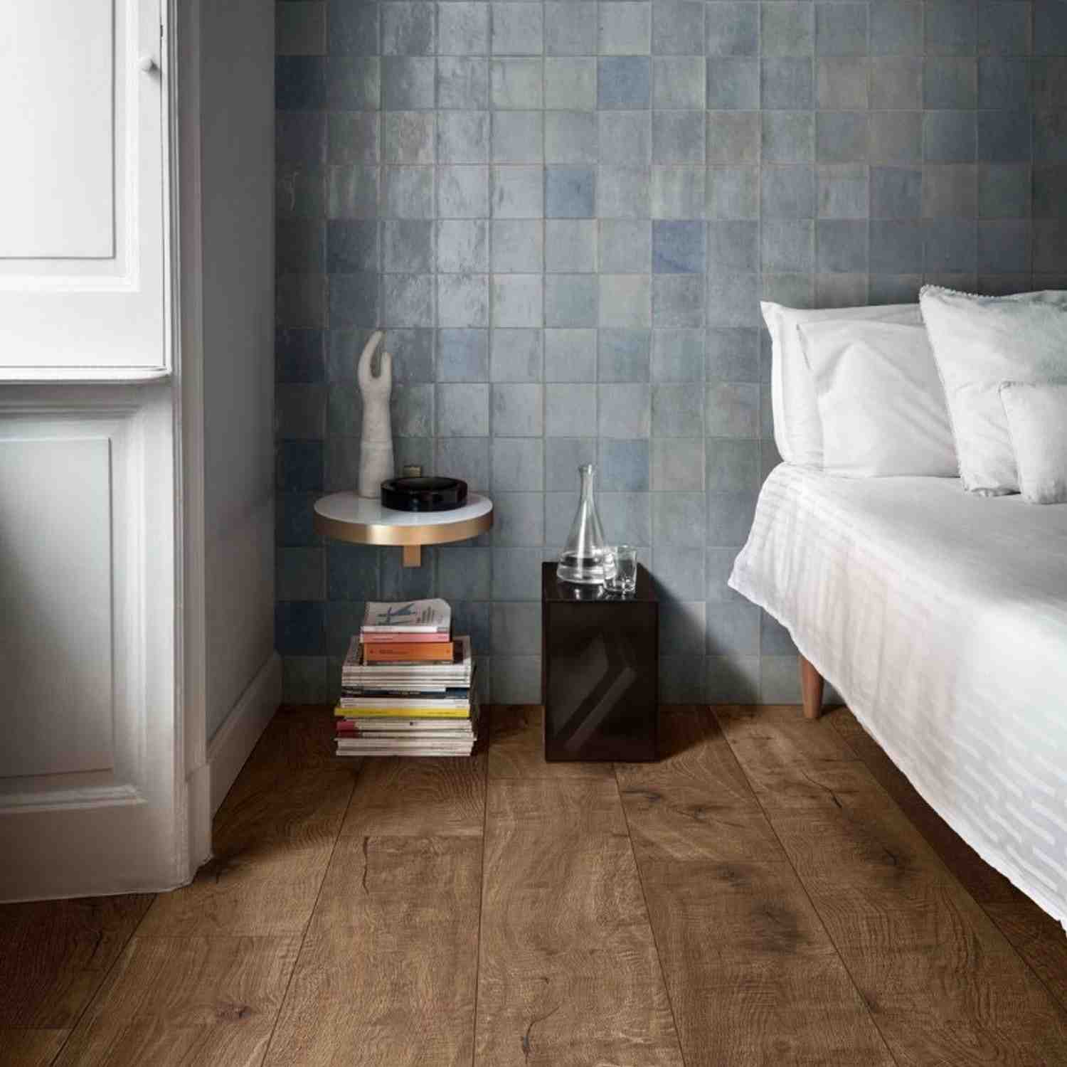 Marazzi Zellige glossy ceramic wall tiles in blue-green tones in bedroom with white bedding, round side table, stacked books, and wooden flooring