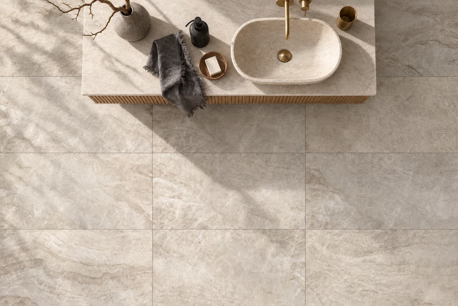 Overhead view of Taj Mahal honed quartzite tile in soft taupe on a bathroom floor, beside a fluted wood vanity with an oval stone basin and brass faucet