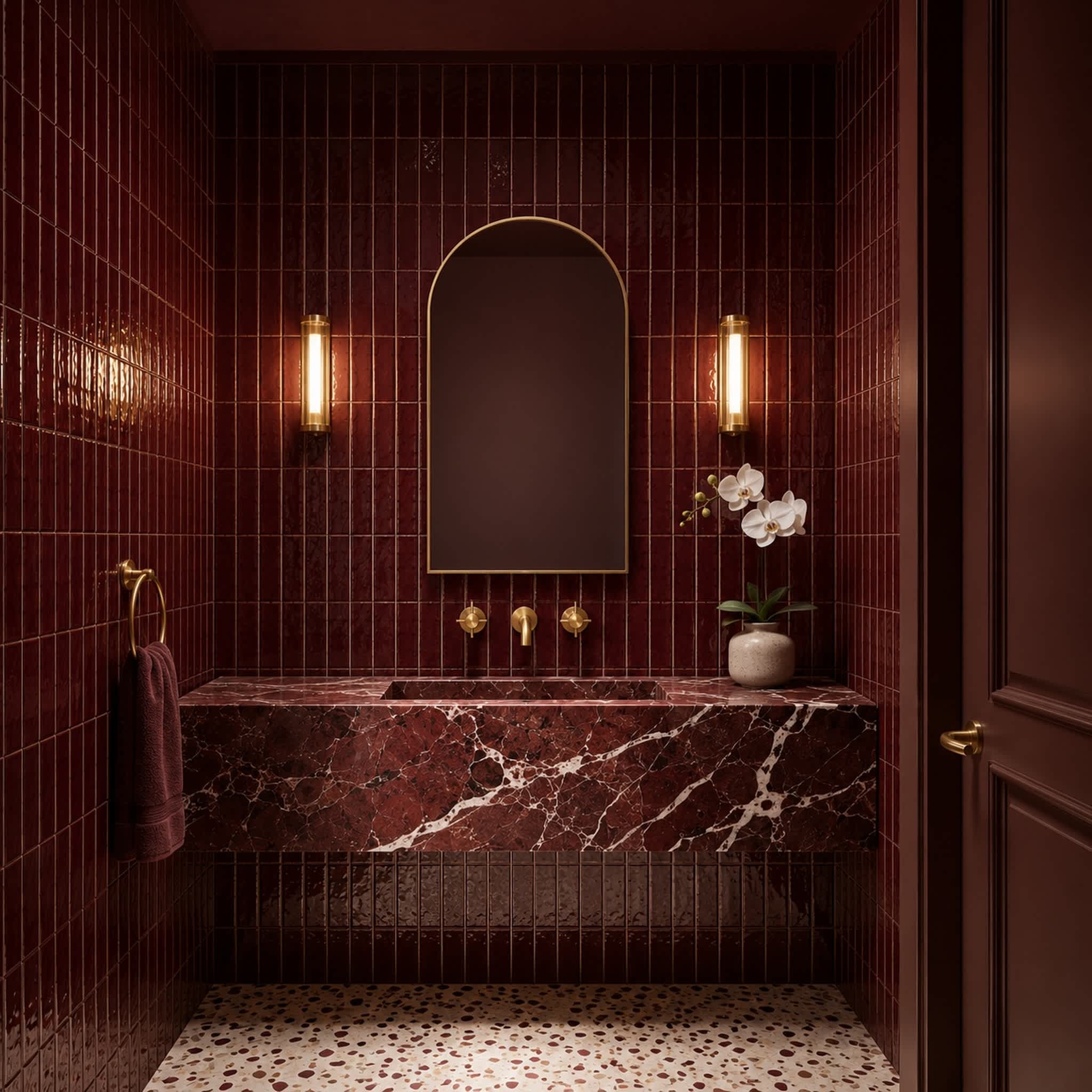 Vivid ceramic tile in Burgundy in a powder room, glossy tiles stacked vertically around an arched brass mirror with a marble sink and terrazzo floor